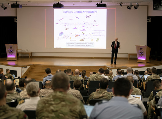 Civilian walking across a stage delivering a presentation to tri-service personnel.