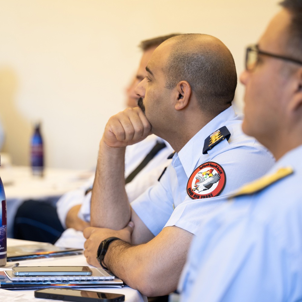 Military officers attentively listening during a briefing in a conference room, with notebooks and water bottles on the table.