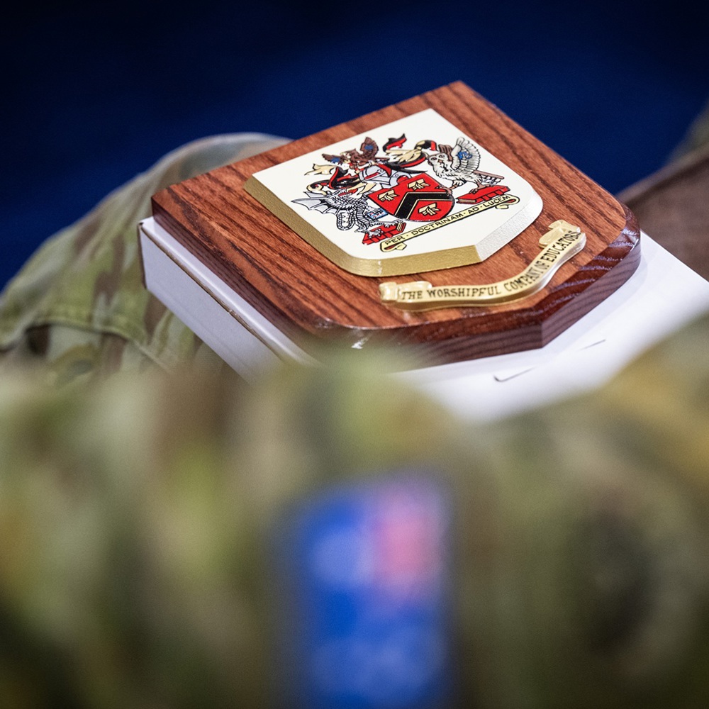 Wooden plaque with a detailed family crest featuring a shield and helmet, resting on a white surface against a blurred military uniform background.