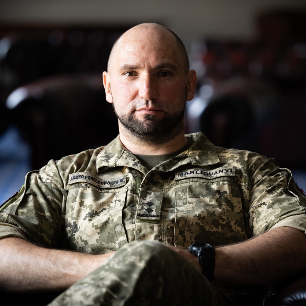 Ukrainian soldier in digital camouflage uniform sitting with arms crossed on a leather chair indoors.