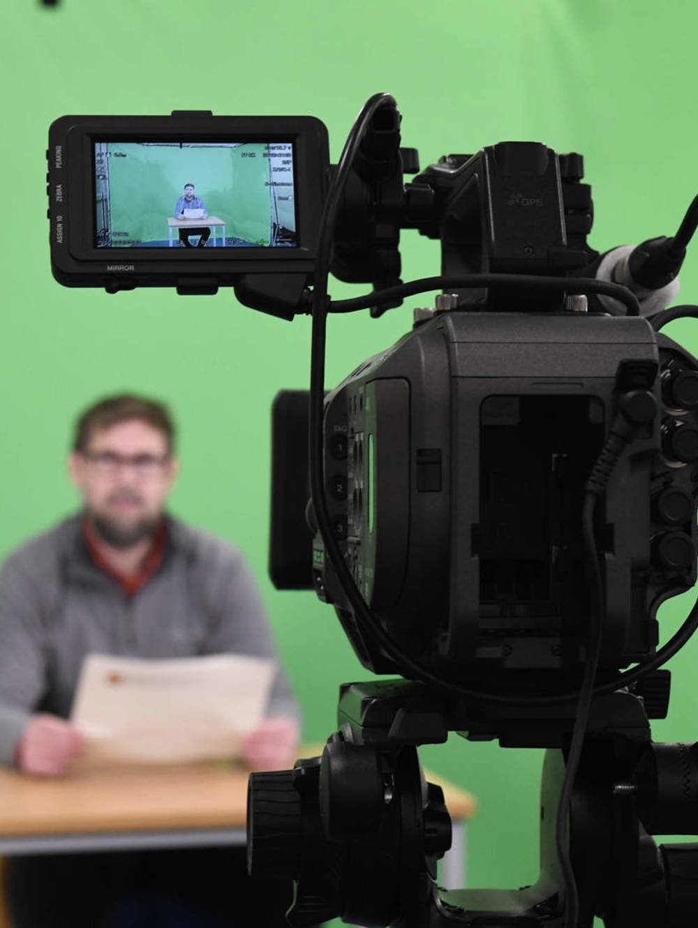 Green screen room with a camera filming a person sat at a desk. 