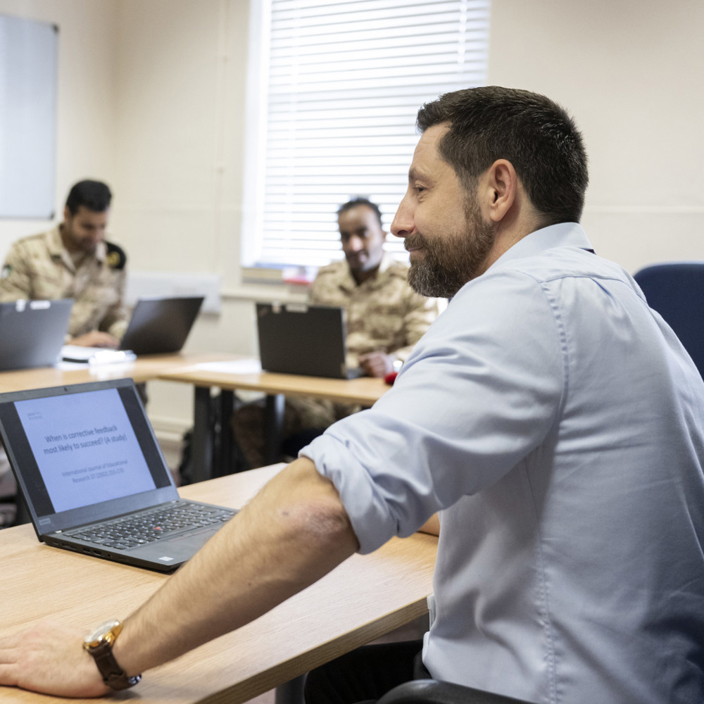 A man dressed in civilian clothes in front of a laptop while teaching Kuwait students. 