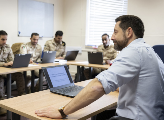 A man dressed in civilian clothes in front of a laptop while teaching Kuwait students. 