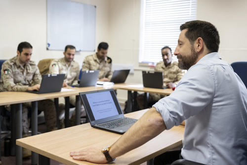 A man dressed in civilian clothes in front of a laptop while teaching Kuwait students. 