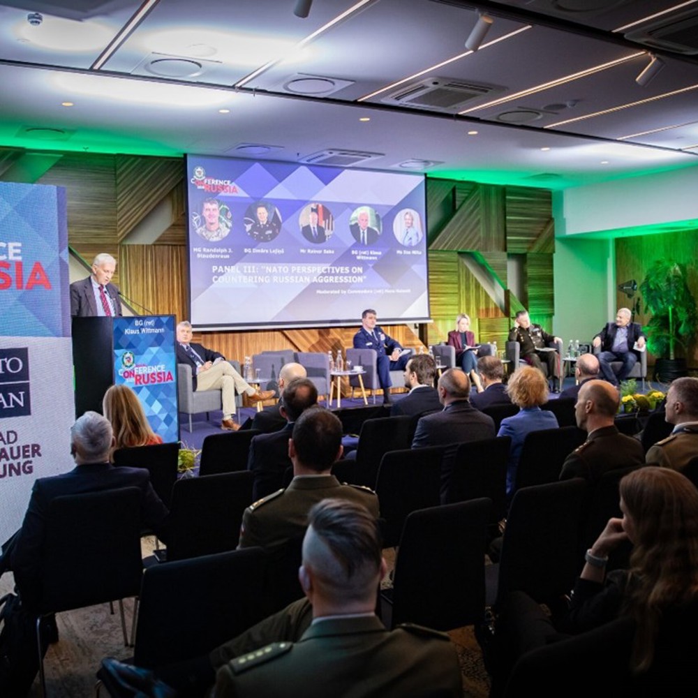 A stage lit by neon lights with a presenter and audience. The stage is flanked by banners showing NATO branding. 