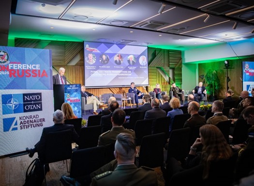 A stage lit by neon lights with a presenter and audience. The stage is flanked by banners showing NATO branding. 