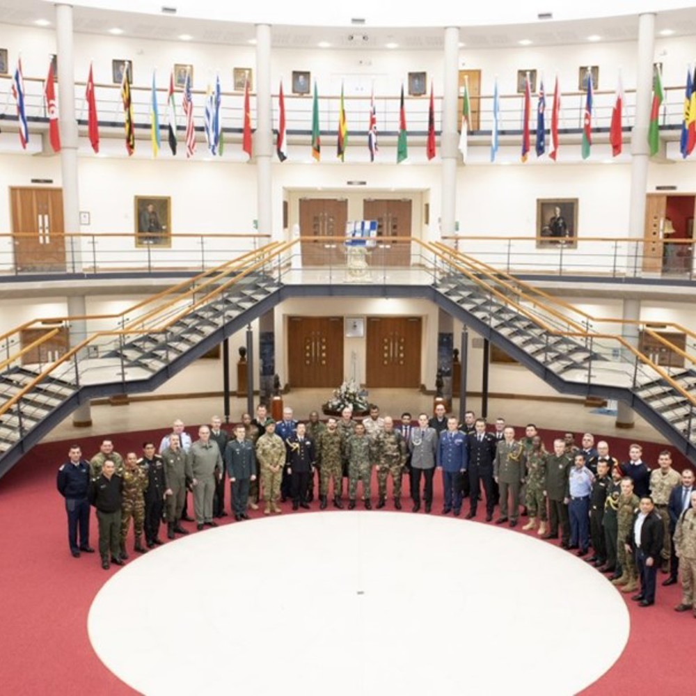 A group of personnel stood in a semi-circle surrounded by overhead international flags. 