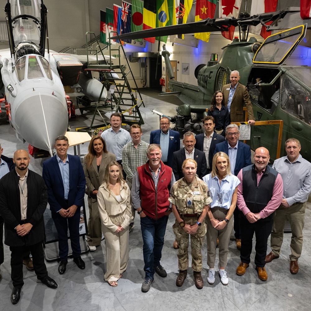 Group of diverse professionals posing inside an aircraft hangar with fighter jets and helicopters in the background.