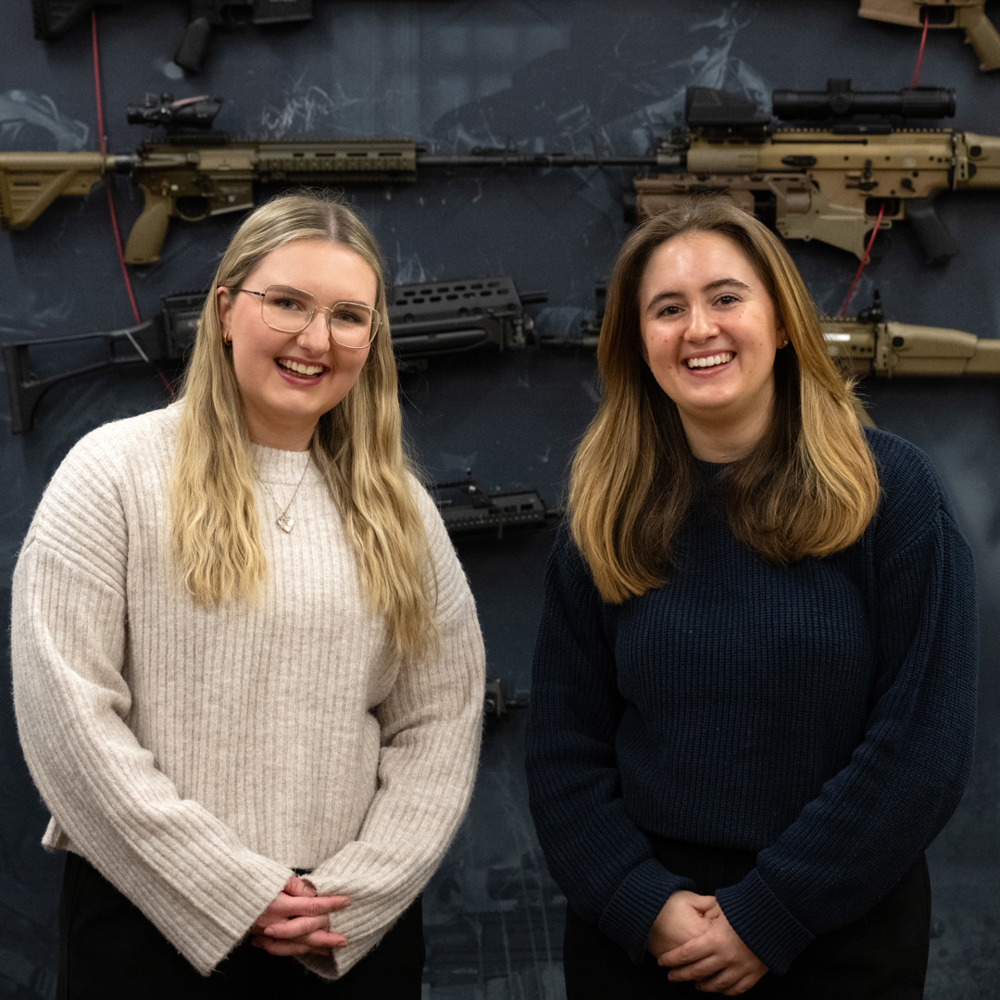 Two young women stood in front of a blue wall with guns, smiling
