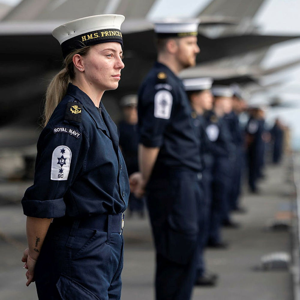 Royal Navy personnel stand in formation on the deck of a naval ship, wearing dark uniforms and white hats labeled HMS Prince.