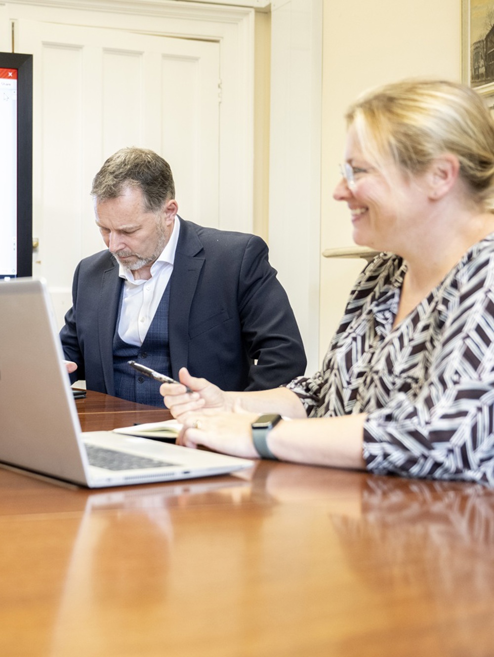 Three people are seated around a wooden conference table, each using a laptop. Behind them, a large screen displays a presentation titled "DLaB Management Board." The room has light-colored walls and a closed white door.