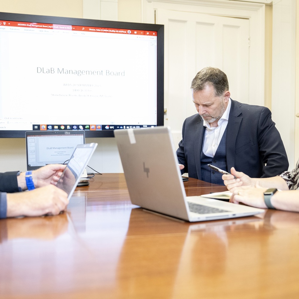 Three people are seated around a wooden conference table, each using a laptop. Behind them, a large screen displays a presentation titled "DLaB Management Board." The room has light-colored walls and a closed white door.