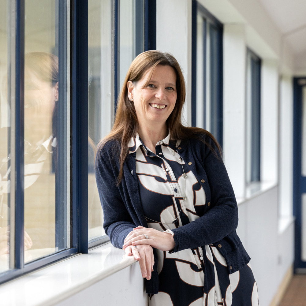 A woman leant against a sill in a long corridor with windows either side. 