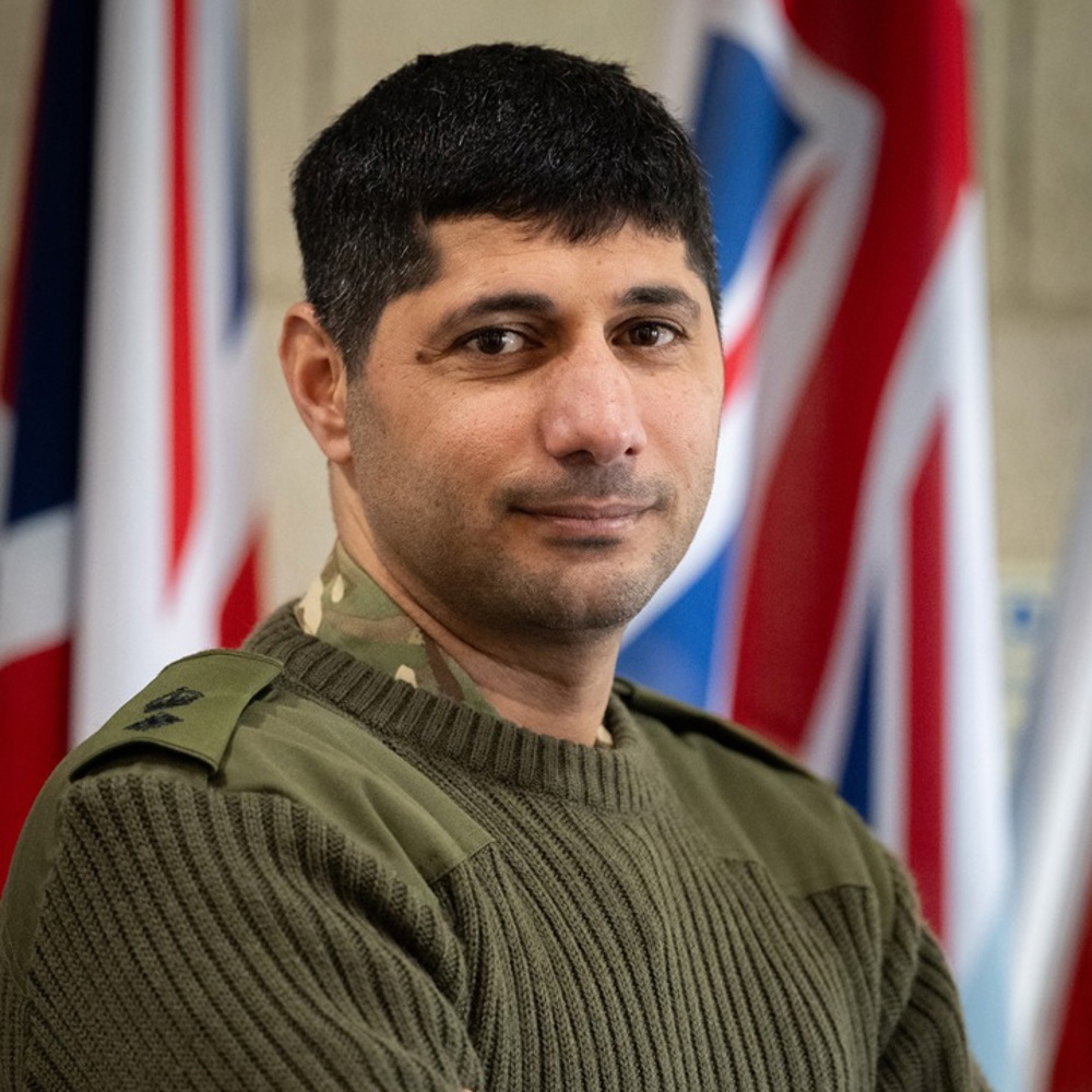 Person in green military sweater with folded arms standing in front of multiple flags indoors.