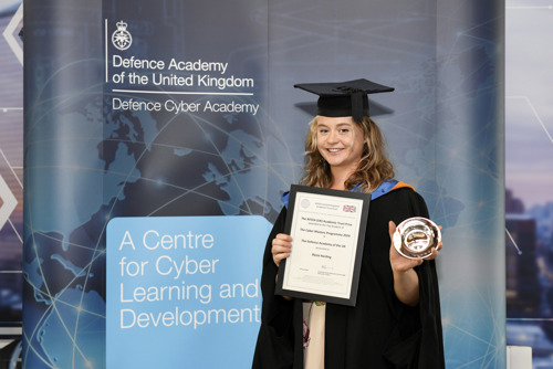 A woman holding a certificate and an award for top student. She is stood in front of a banner advertising the Defence Cyber Academy
