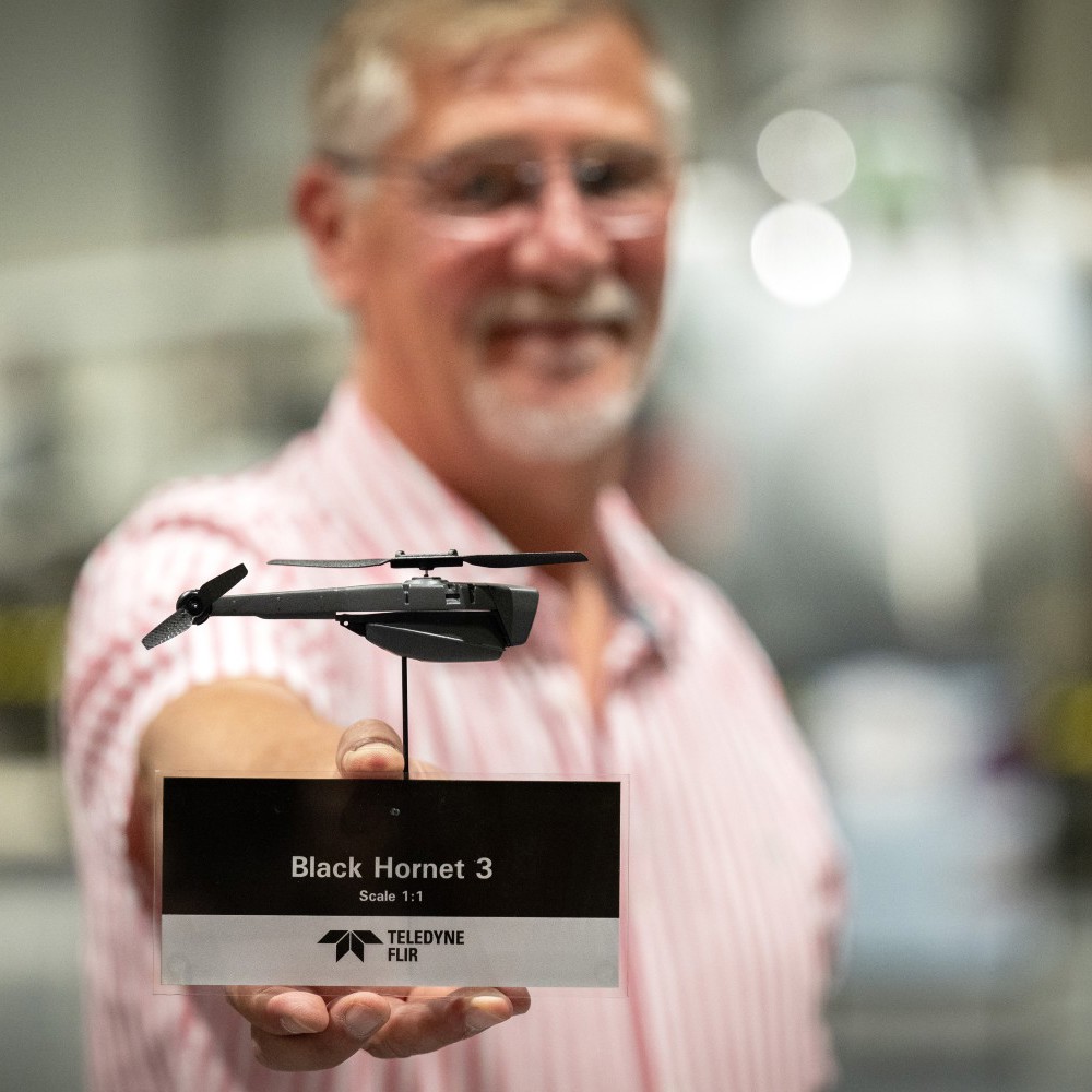A man (out of focus) holding a very small black drone shaped like a helicopter.