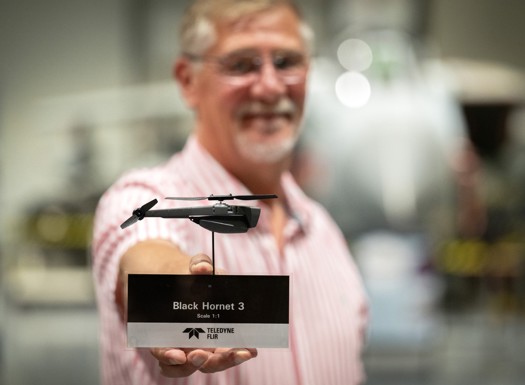 A man (out of focus) holding a very small black drone shaped like a helicopter.