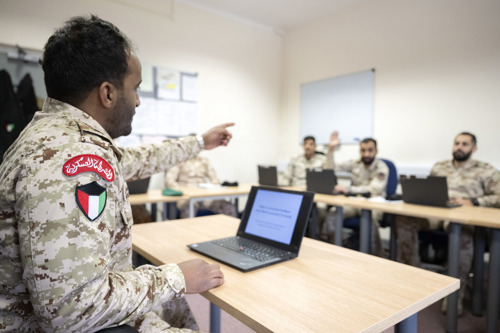 A man dressed in Kuwait military uniform in front of a laptop while teaching Kuwait students. 
