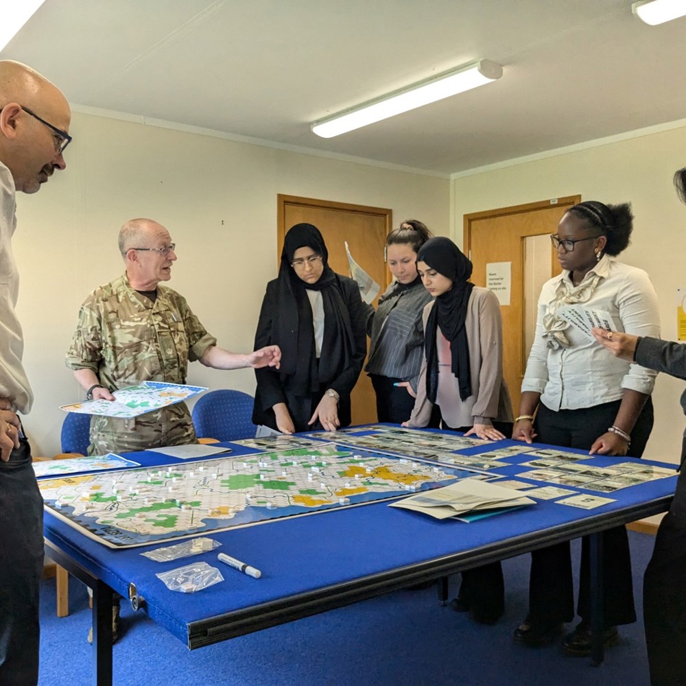 Students and MOD personnel in a classroom, wargaming around a table. 