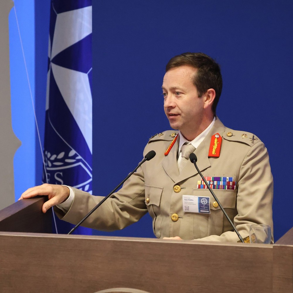 Military officer in uniform speaking at a podium with a NATO flag in the background.