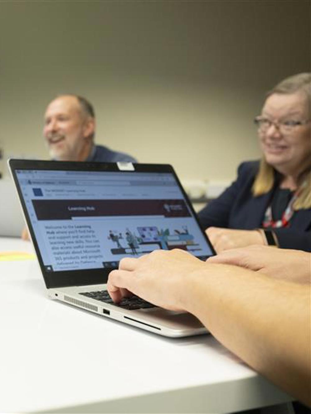 Hands typing on a laptop during a meeting with colleagues seated around a white conference table and a presentation screen in the background.