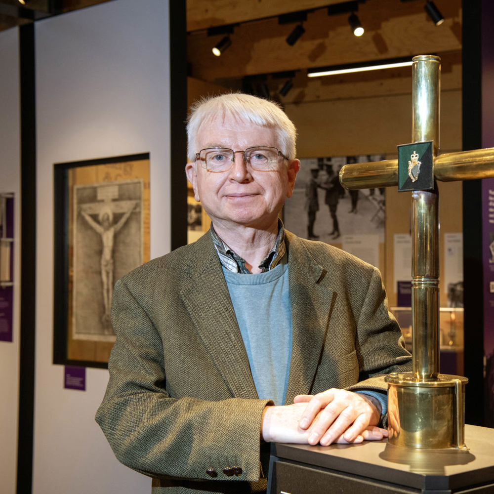 A man stood in a military museum stood next to a gold cross exhibit with faith in forces displays in the background. 
