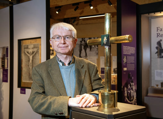 A man stood in a military museum stood next to a gold cross exhibit with faith in forces displays in the background. 