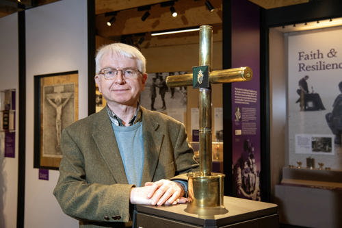 A man stood in a military museum stood next to a gold cross exhibit with faith in forces displays in the background. 