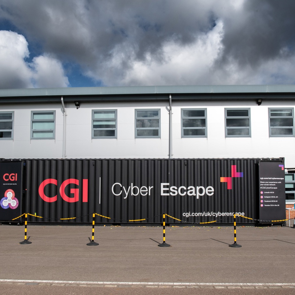 Black shipping container with CGI Cyber Escape branding outside a white building under a cloudy sky.