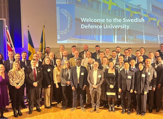 Group photo of diverse professionals standing on stage at the Swedish Defence University event with flags and a welcome message in the background.
