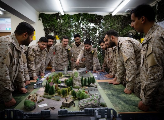 A group of international officers stood around a table with a wargaming boardgame in the centre. The room is decorated with fake camouflage filigree. 