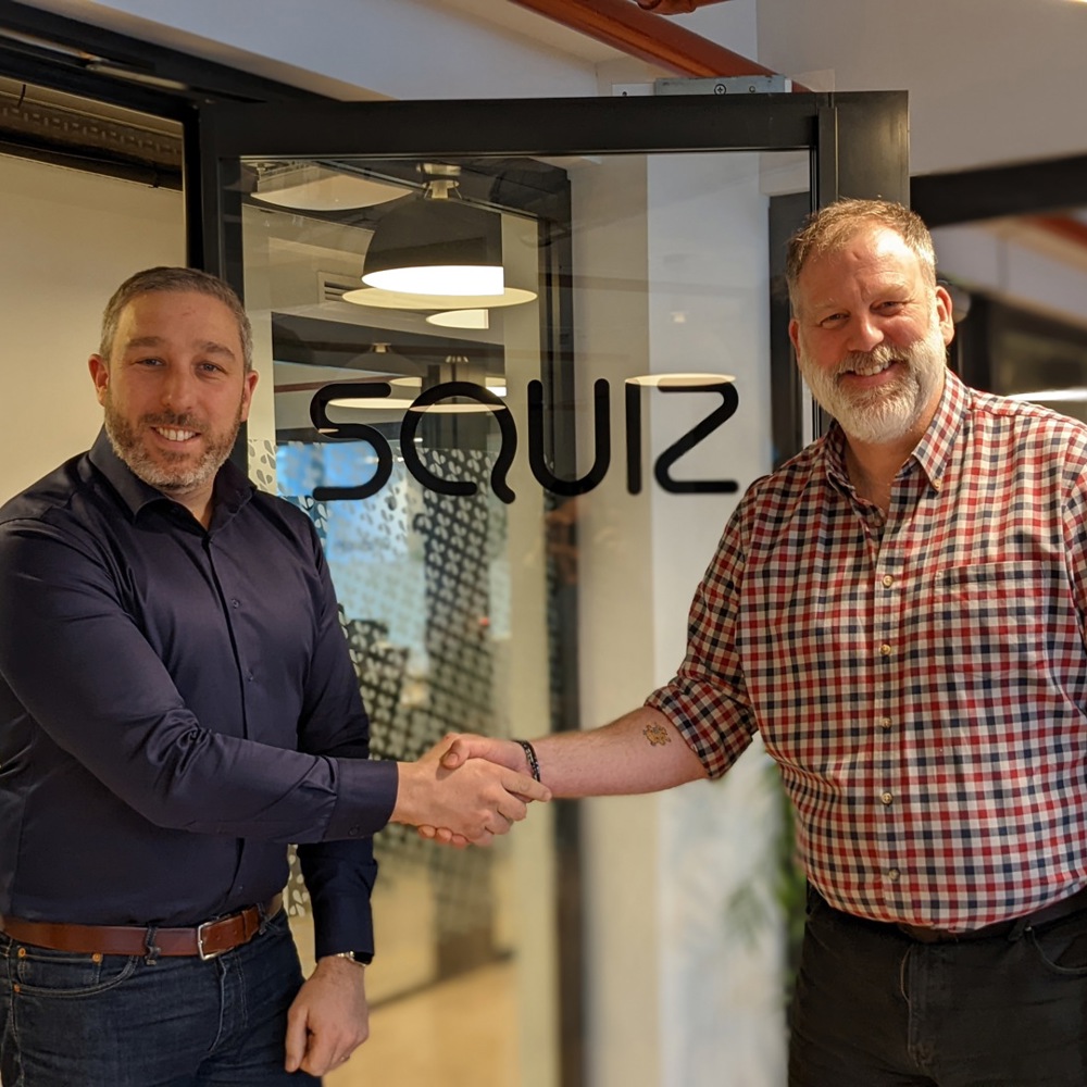 Two men in civilian dress shaking hands in front of a glass door with the company name 'Squiz'.