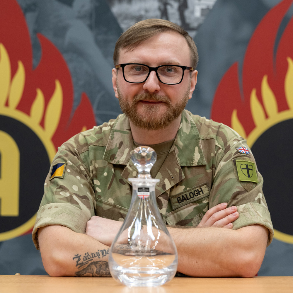An army officer wearing glasses sat with glass decanter award in front of him. 