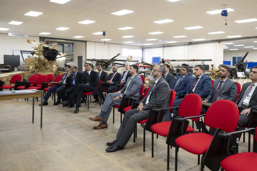 Pakistan delegation listening to a presentation seated in a large hall with military equipment in the background. 