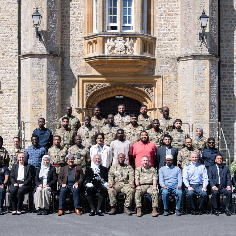 Large group of military personnel and civilians posing in front of a historic stone building with arched entrance and windows.