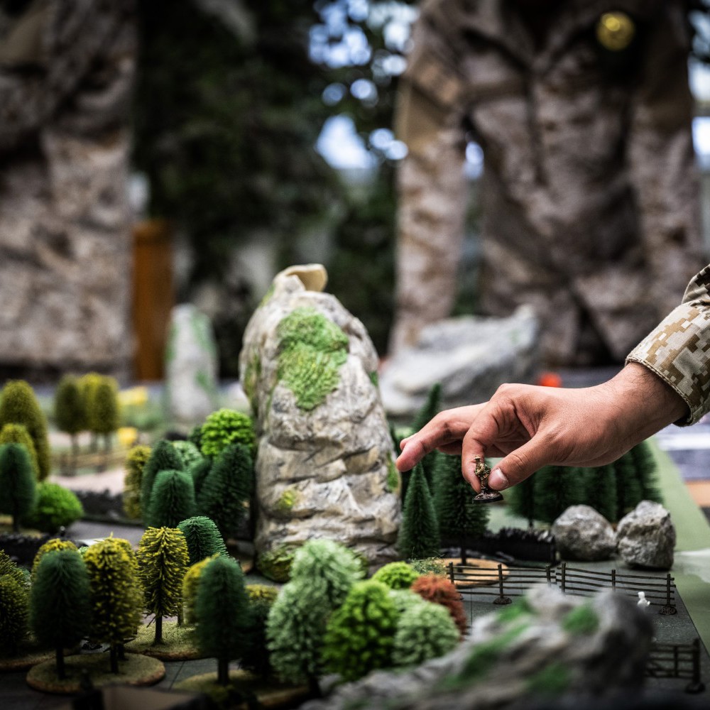 A close up of a wargaming board with model trees, rocks and fencing with a hand picking up a model soldier. 