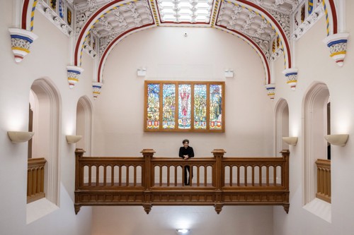 Female Reverand standing on an internal balcony with religious coloured glass pane windows behind her.