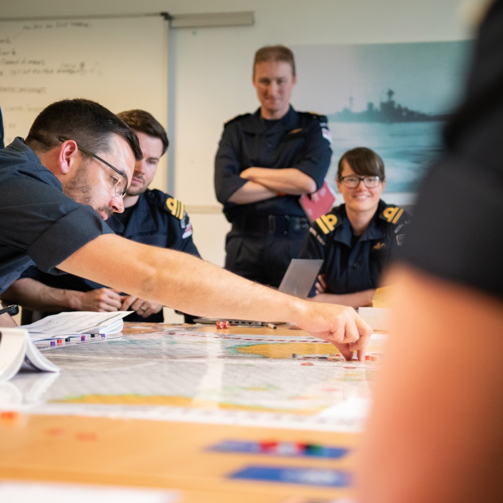 Group of naval officers in uniform gathered around a table studying maps and documents during a strategy meeting.
