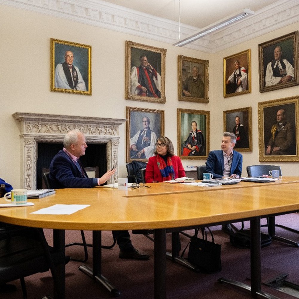 People sat around a table having a discussion surrounded by portrait oil paintings on the walls. 