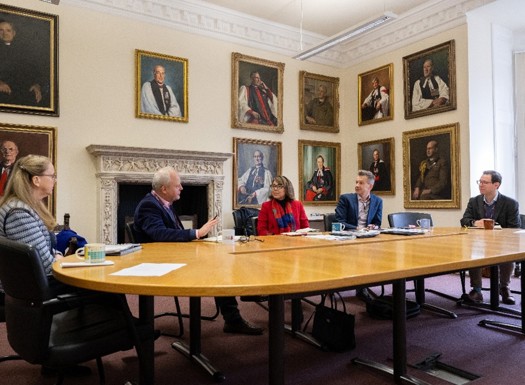 People sat around a table having a discussion surrounded by portrait oil paintings on the walls. 