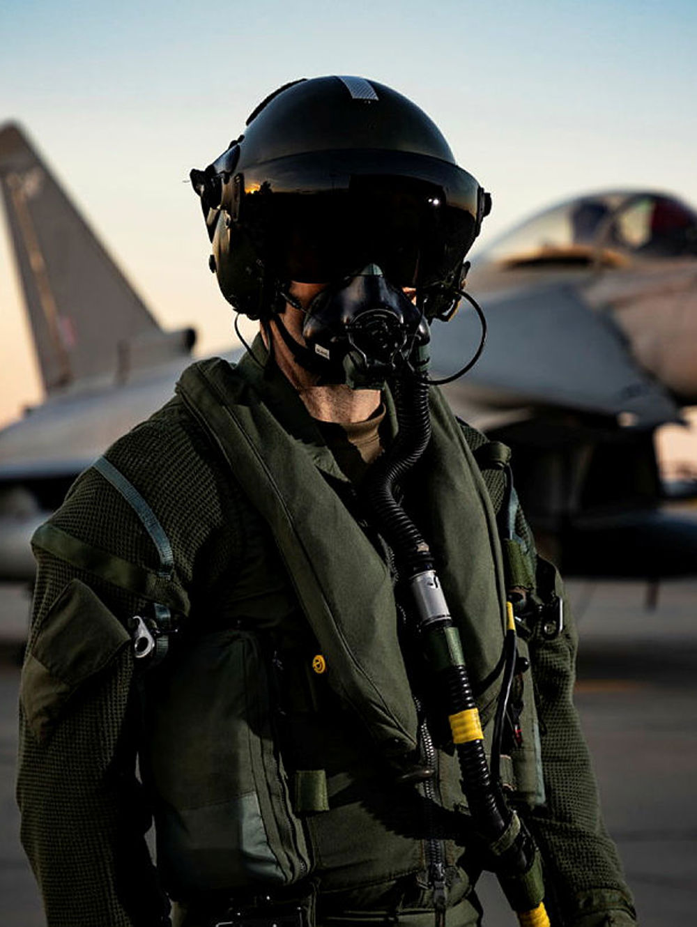 Military pilot in full flight gear and helmet standing on the tarmac with a fighter jet in the background at sunset.