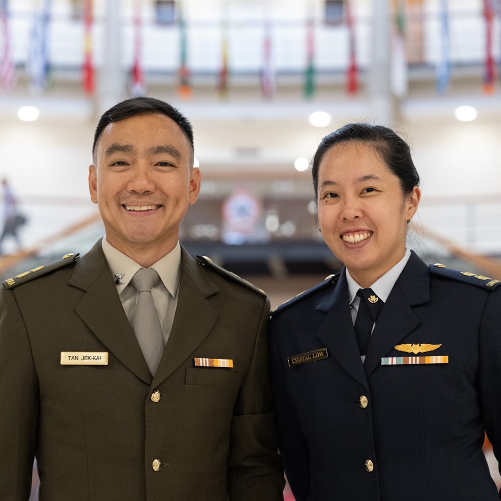 Two smiling military officers in uniform standing side by side inside a large building with flags hanging above.