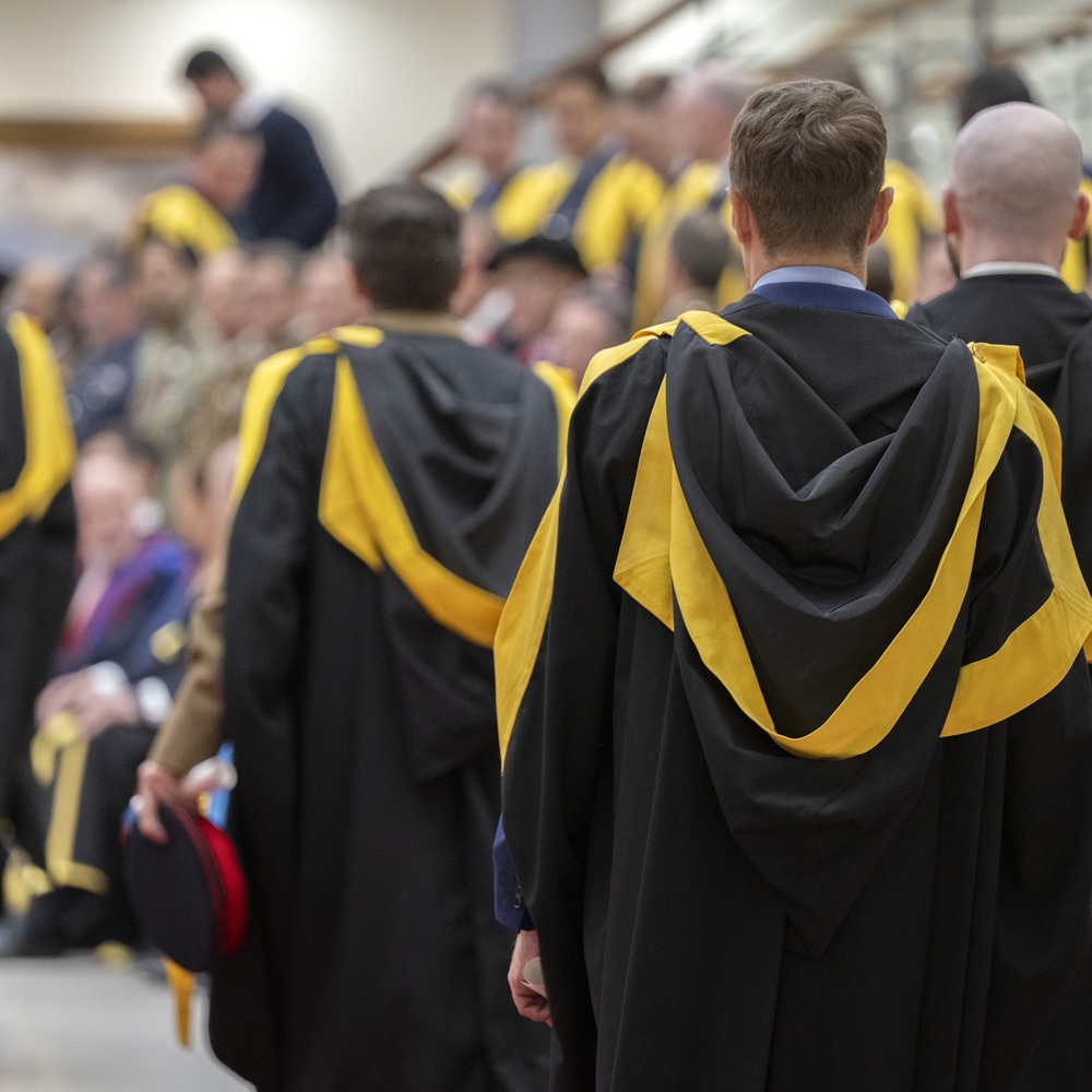 Group of graduates in black gowns with yellow hoods gathered indoors, preparing for a ceremony.