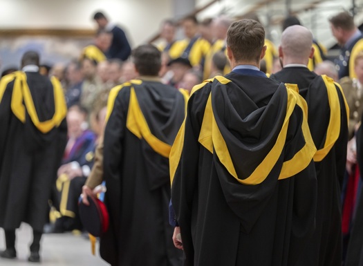 Group of graduates in black gowns with yellow hoods gathered indoors, preparing for a ceremony.