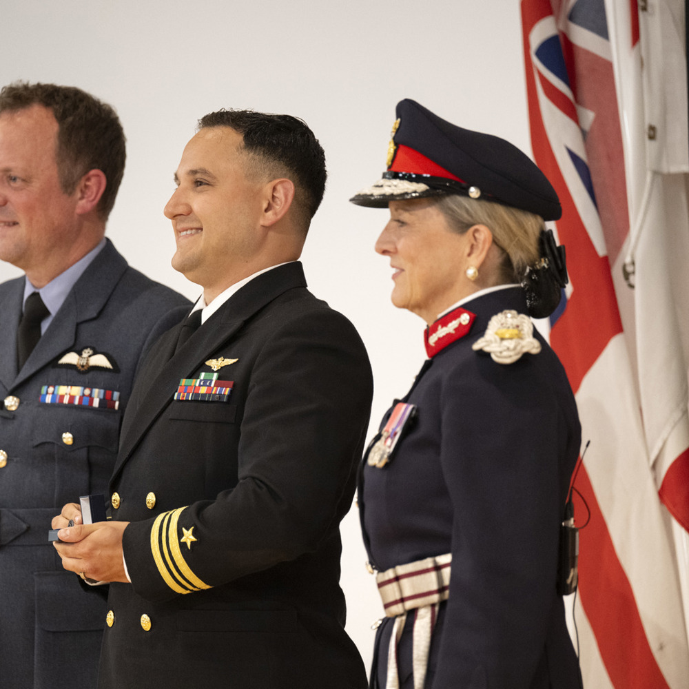Three people in uniform with military flags in the background.