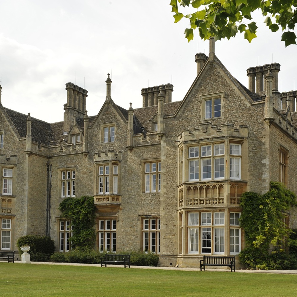 A large, historic stone mansion with multiple chimneys, pointed gables, and tall windows. It is surrounded by manicured lawns, greenery, and benches. Tree branches with green leaves frame the top of the image under a partly cloudy sky.