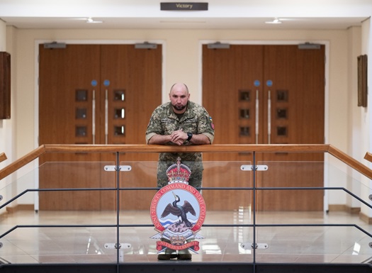 Military personnel in camouflage uniform leaning on a glass railing inside a building with wooden double doors behind.