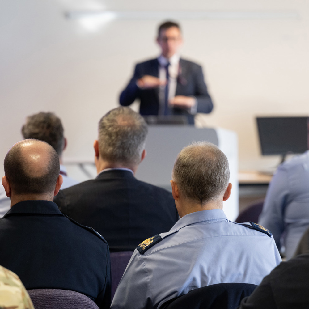 A man in a suit presenting to a diverse group of civilians and military personnel.