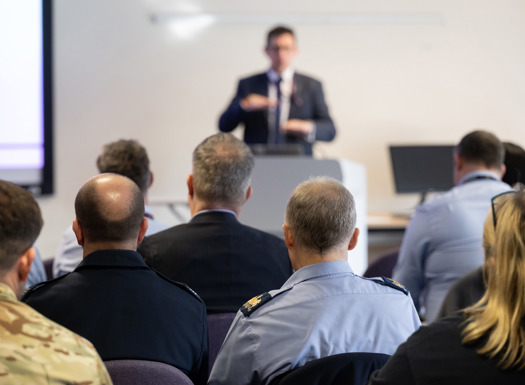 A man in a suit presenting to a diverse group of civilians and military personnel.