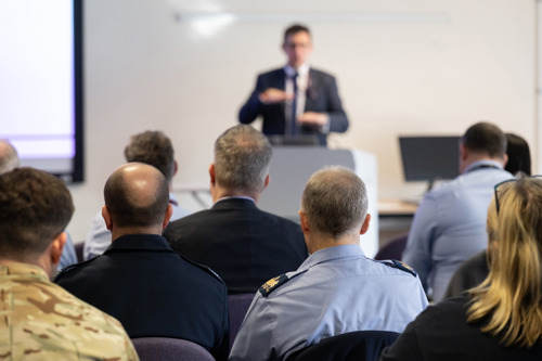 A man in a suit presenting to a diverse group of civilians and military personnel.